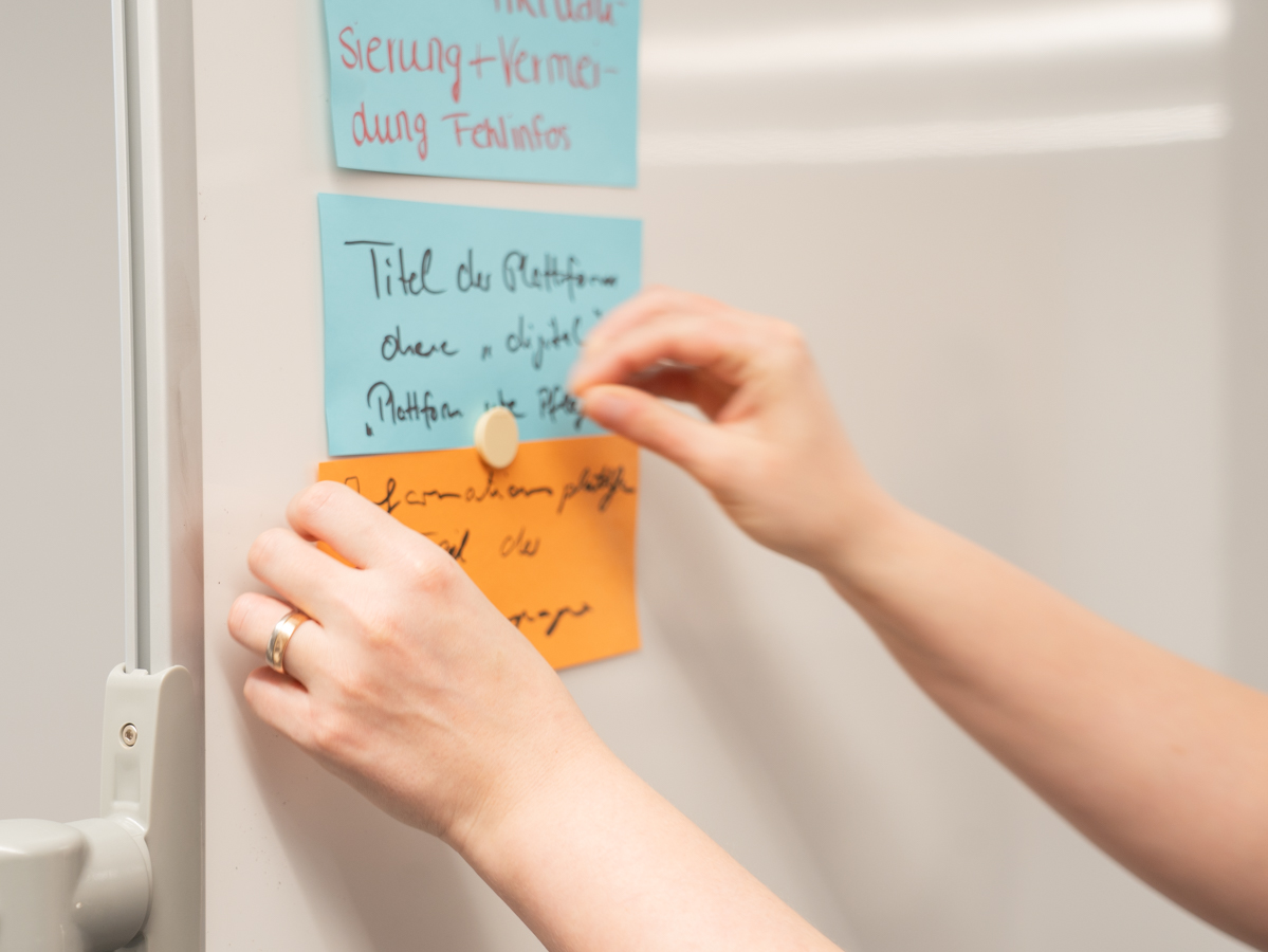 Zwei Hände hängen mit einem Magnet ausgefüllte Moderationskarten an ein Whiteboard