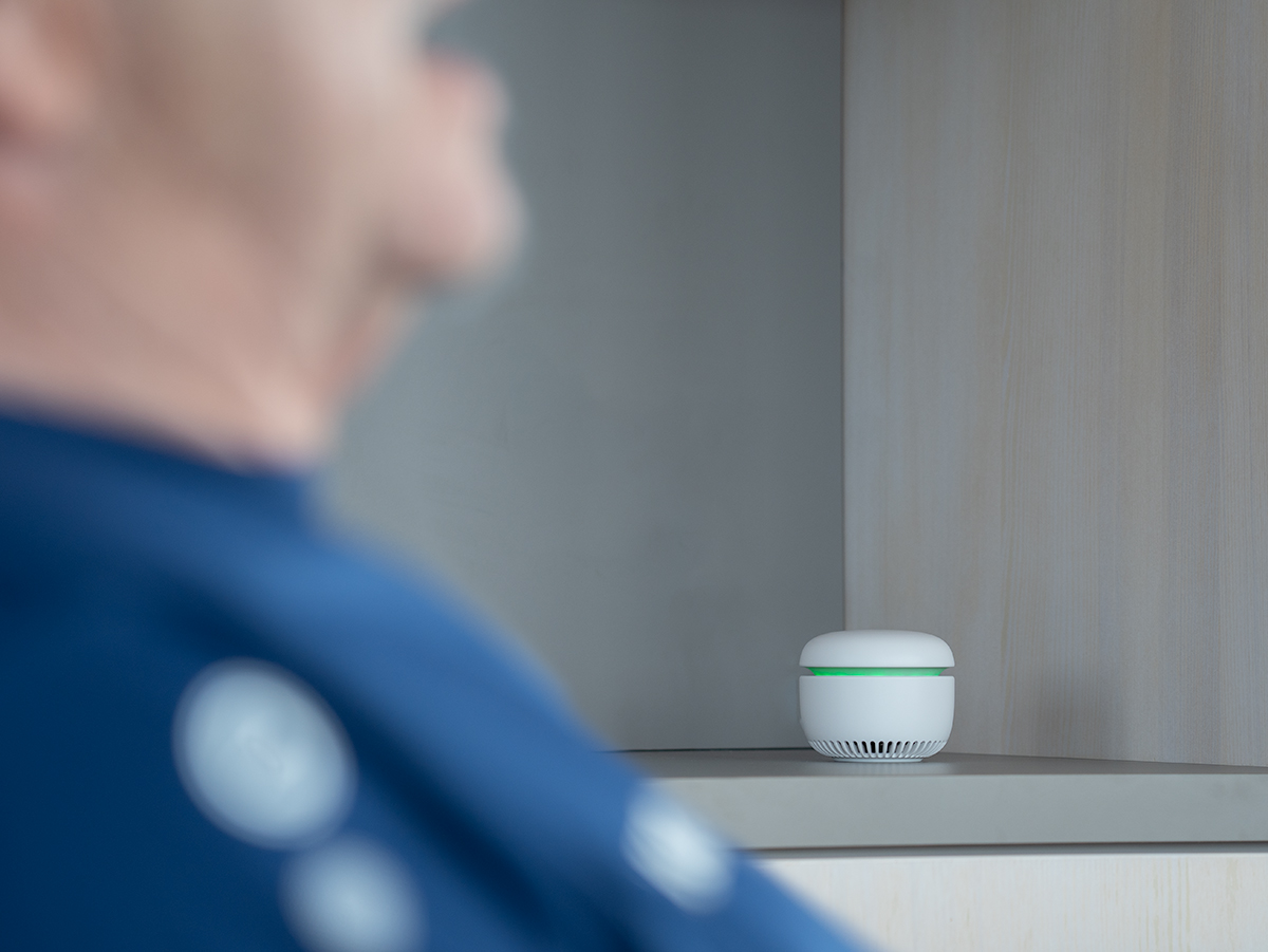 Im unscharfen Vordergrund spricht ein Senior. Im scharfen Hintergrund steht ein Hausnotruf, der grün leuchtet.