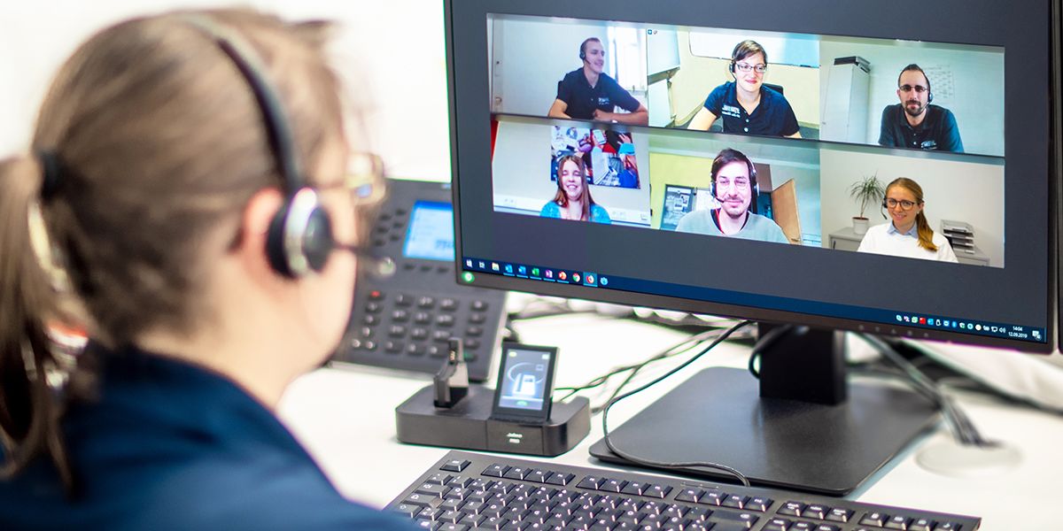 Eine Frau sitzt vor einem Monitor. Auf dem Monitor sind weitere Personen in einer Videokonferenz zu sehen.
