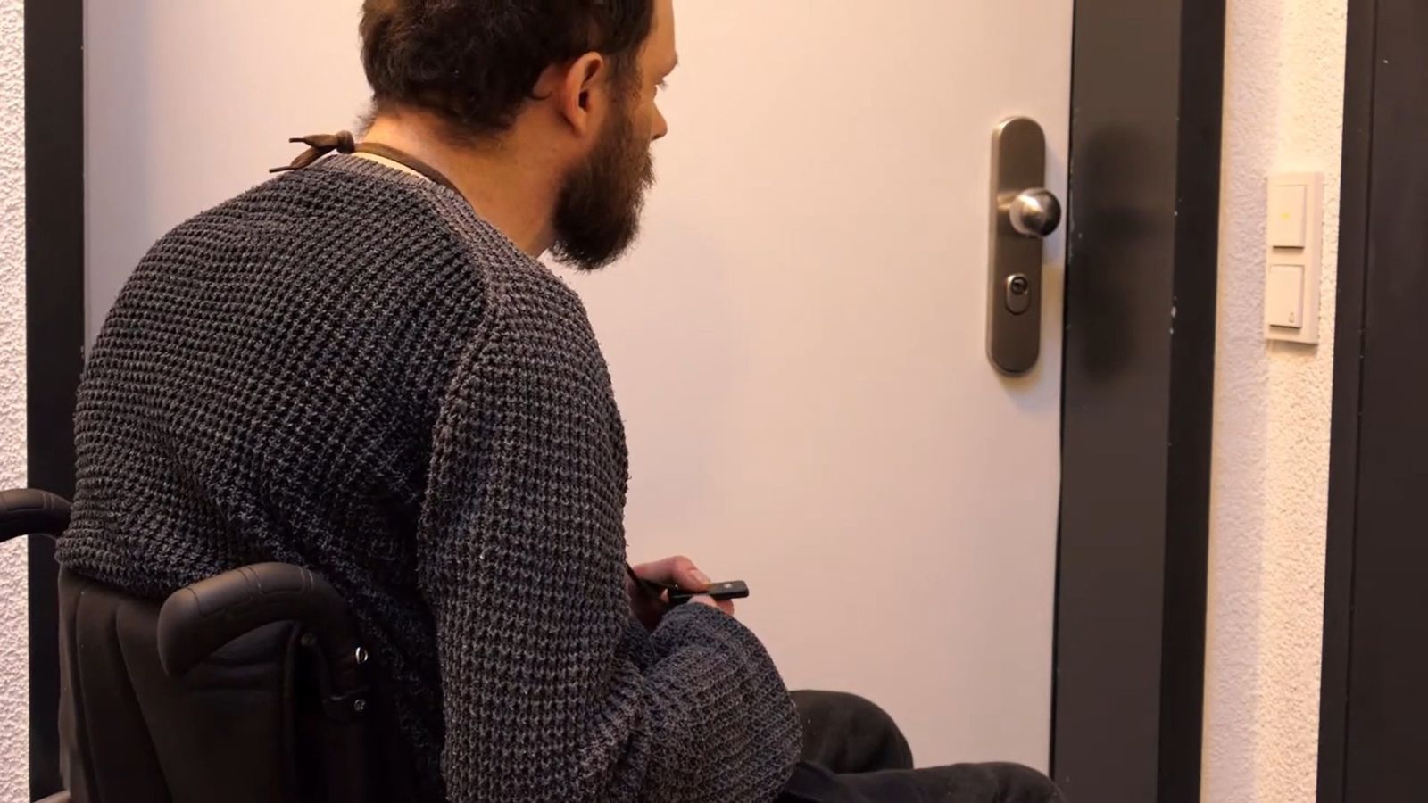 Ein Mann im Rollstuhl befindet sich vor einer verschlossenen Wohnungstür und hält eine Fernbedienung in der Hand.