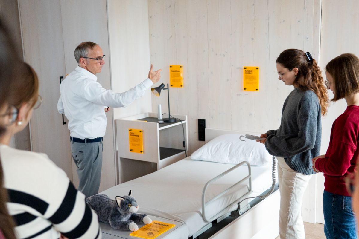 Junge Personen stehen vor einem Pflegebett und ein Mann erklärt das Pflegebett.