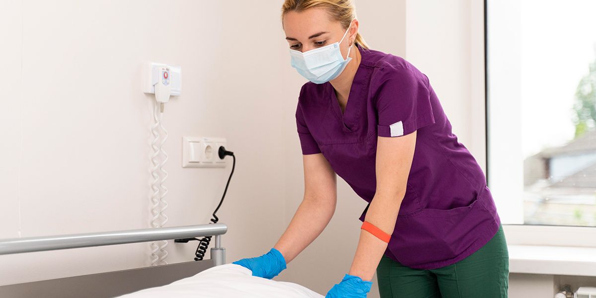 Eine Pflegefachperson mit medizinischer Maske und Hygienehandschuhen richtet das Kopfkissen auf einem Pflegebett.