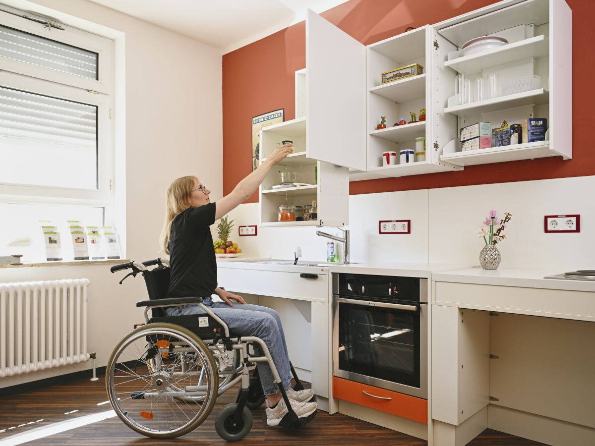 Weibliche Person im Rollstuhl entnimmt Gegenstand aus Hochschrank der barrierefreien Küche in der Musterwohnung