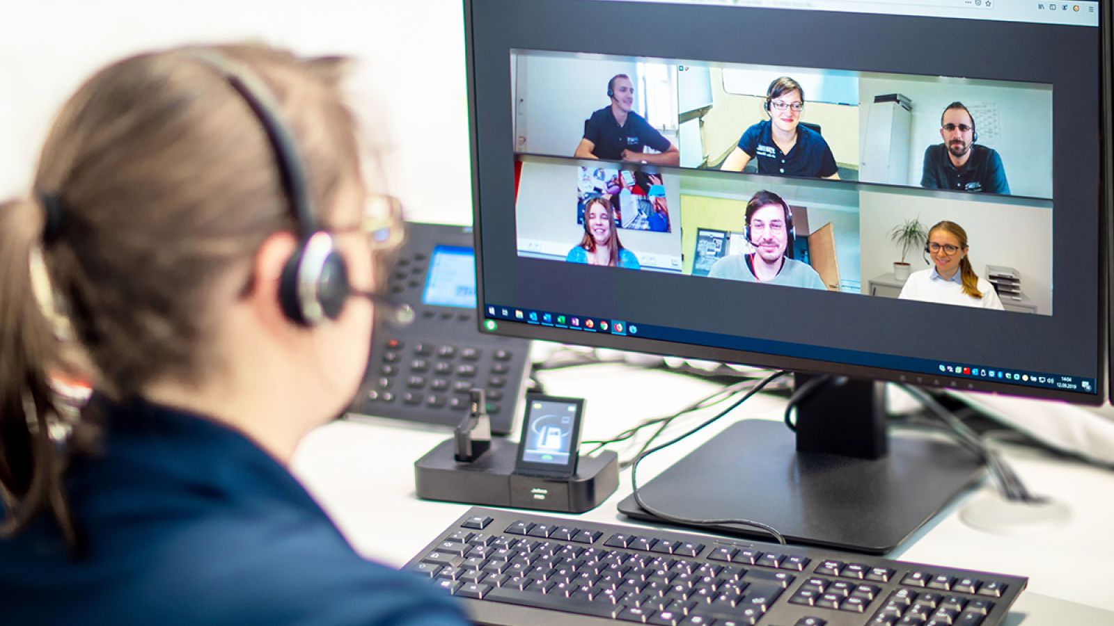 Eine Frau sitzt vor einem Monitor. Auf dem Monitor sind weitere Personen in einer Videokonferenz zu sehen.
