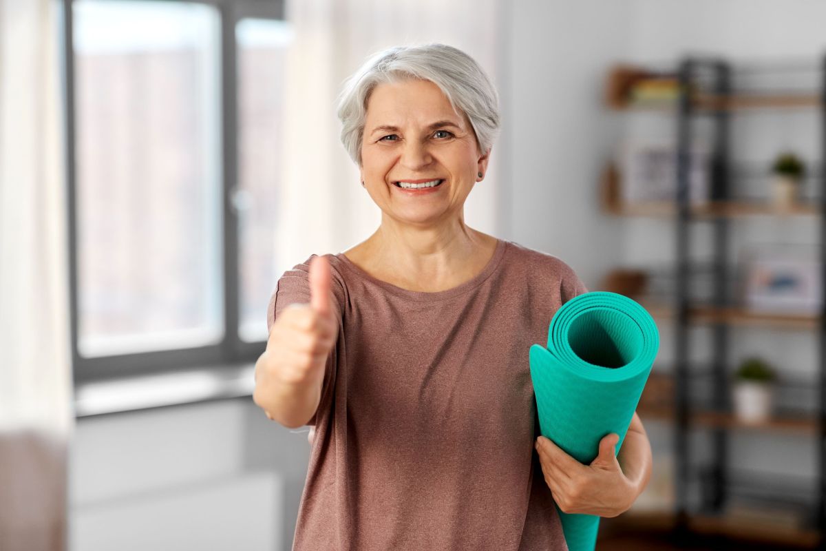 Eine Seniorin hält eine zusammengerollte Gymnastikmatte in den Armen und hält einen Daumen hoch.